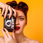 Portrait of amazed pinup woman with camera. Charming photographer with red lips taking pictures.